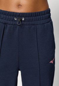 Mornarsko modri trenirki z vrvico v pasu, ki imata stranske žepke in roza logotip. Material je mehak in gladke teksture.