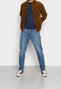 Chaqueta de pana marrón sobre una camiseta de color azul marino, combinada con jeans de un azul claro y zapatillas marrones. Ajuste casual con detalles de costura visibles.