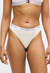 Beige string veļa ar baltu elastīgu jostu, kas rotāta ar "TOMMY HILFIGER" logo tumši z blue un sarkanas krāsas. Gluda tekstūra, piegulošs dizains.