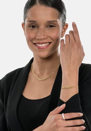 Gold chain necklace and bracelet with linked designs, set against a black outfit. Smooth texture, shiny finish, worn on a model's neck and wrist.