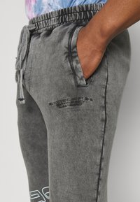 Šedé tepláky vyrobené z měkké, strukturované látky. Mají elastický pas, boční kapsy a potištěný vzor na nohavici.