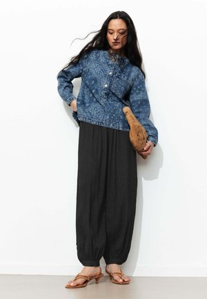Femme portant une veste en jean bleue à motifs, un pantalon noir ample, des sandales à talons beiges, tenant une pochette en cuir beige sur fond blanc.