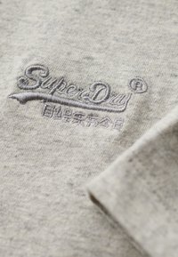 Tessuto di cotone grigio con logo ricamato "SuperDry" in grigio scuro. Texture morbida con un motivo melange. Il dettaglio mostra chiaramente le cuciture.