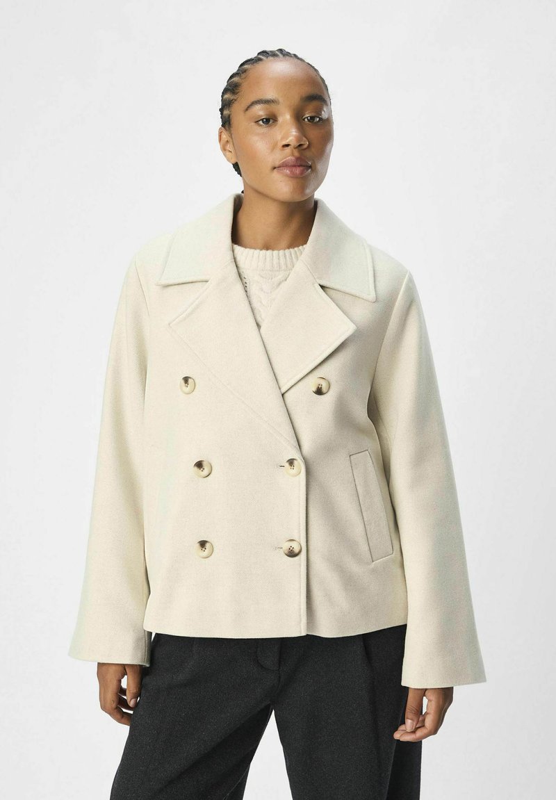 Veste double boutonnage beige clair avec un large col, de grands boutons ronds et des poches latérales ; tissu texturé et coupe décontractée.