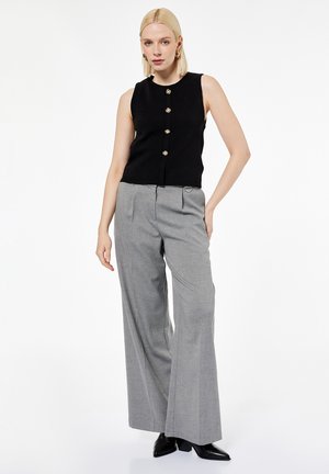 Femme portant un haut noir sans manches boutonné, un pantalon gris taille haute à jambes larges, et des chaussures noires à bout pointu, debout avec la main sur la hanche.