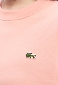 Κοντινό πλάνο ροζ υφάσματος με μικρό πράσινο λογότυπο κροκοδείλου κεντημένο κοντά στη γραμμή του λαιμού.