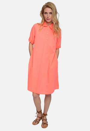 Blonde woman wearing a loose coral shirt dress with short sleeves and tan lace-up sandals, standing with hands in pockets.