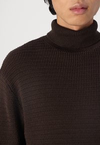 Pull à col roulé marron avec un motif texturé en point gaufré. Le design présente un col haut et une coupe ample pour le confort.