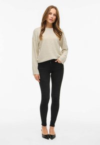 Ženska z dolgimi svetlo rjavimi lasmi oblečena v bež pulover, črne ozke kavbojke in črne usnjene petke, stoji ob beli steni.