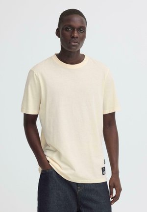 Tume nahaga mees, kes kannab lihtsat heledat kreemjat lühikese varrukaga T-särki ning tumedat sinist teksapükse, seisab ühetaustase heleda tausta ees.