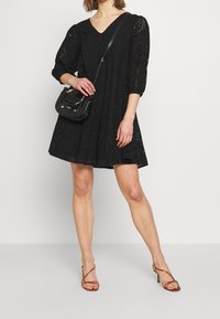 Musta mekko V-pääntiellä, läpikuultavat puffihihat ja silkkipitsikuvio. Pariutettu mustan olkalaukun ja ruskeiden remmissandaalien kanssa.