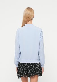 Veste bomber bleu clair avec col et poignets côtelés, associée à une jupe noire ornée d'un motif floral. Texture lisse, design décontracté.