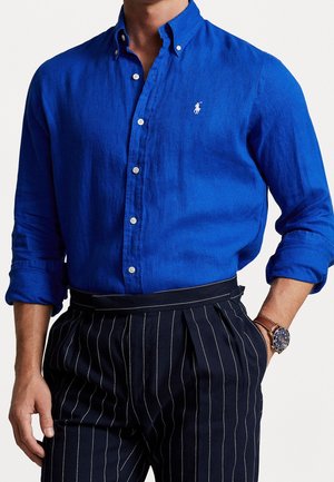 Homme portant une chemise bleue royale à boutons avec manches retroussées, un pantalon noir à fines rayures et une montre-bracelet avec un bracelet en cuir marron.