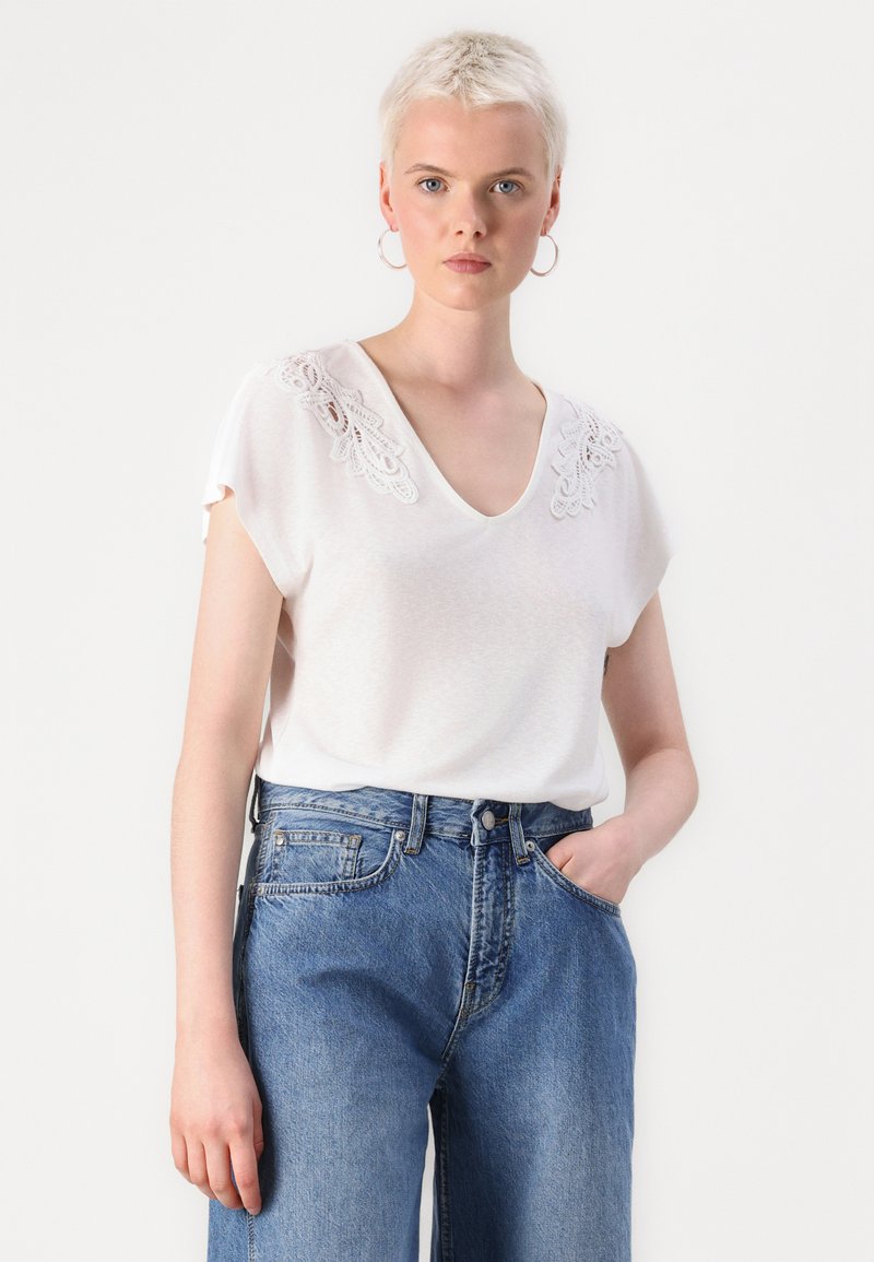 Haut blanc à manches courtes avec un col en V et des détails en dentelle sur les épaules, associé à un jean en denim bleu taille haute. Texture de tissu lisse.