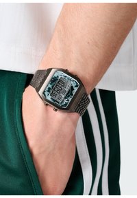 Montre numérique avec un bracelet en métal argenté et un cadran rectangulaire. Écran noir et turquoise avec des boutons sur le côté.