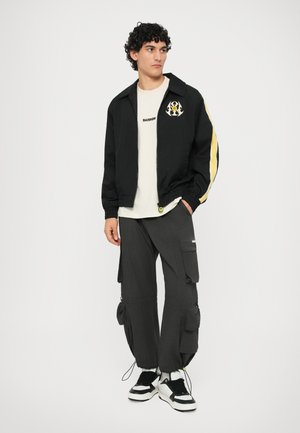 Giubbotto bomber nero con strisce gialle sui lati e un logo, abbinato a pantaloni cargo grigi con più tasche. Sneakers bianche e nere.