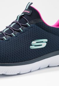 Námořnicky modrá sportovní obuv z síťoviny s tyrkysovým logem "S", růžovým vnitřním lemem, pruhovanými tkaničkami a bílou texturovanou podrážkou.