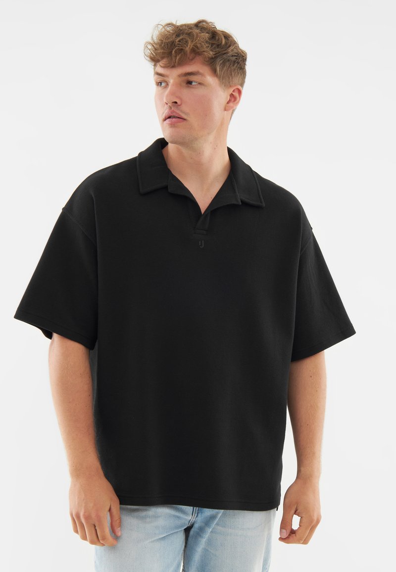 Fiatal férfi göndör hajjal, laza fekete rövid ujjú pólót és világoskék farmernadrágot viselve, balra néz egy fehér háttér előtt.