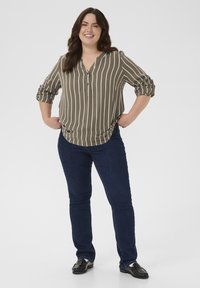 Smiling woman standing with hands on hips, wearing a green striped blouse, dark jeans, and black loafers against a plain background.