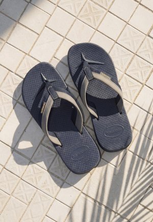 Et par mørkeblå og beige Havaianas flip-flops som hviler på et lyst flislagt gulv med mønster og skyggene av en plante.