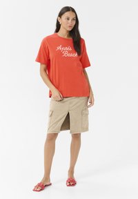 Rød bomulds t-shirt med teksten "Après Beach", styles med en lysebrun denimnederdel med cargolommer og en frontslids; båret med lyserøde blomster sandaler.