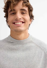 Joven con cabello rizado castaño que lleva un suéter gris claro, sonriendo frente a un fondo blanco liso.