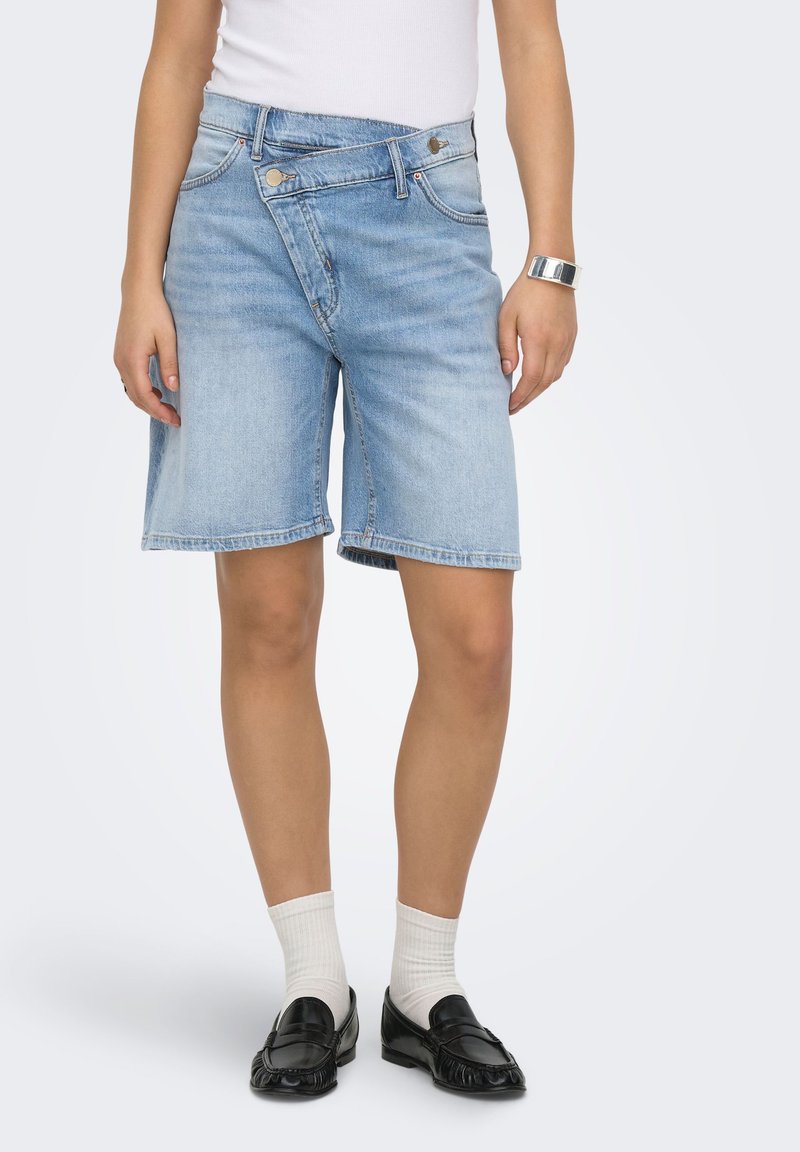 Personne portant un short en jean bleu clair avec une taille boutonnée chevauchante, des chaussettes blanches, des mocassins noirs et un haut blanc sans manches.