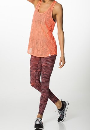 Mujer con una camiseta sin mangas de malla coral, leggings morados con estampado y zapatillas negras para correr, de pie con una pierna cruzada sobre la otra.