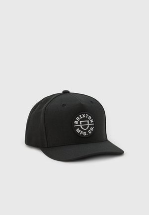 Casquette de baseball noire avec un patch rond à l'avant présentant le texte "BRIXTON MFG. CO." et un logo de bouclier stylisé en broderie blanche.