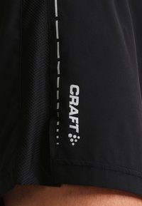 Närbild av svarta idrottsshorts med meshpanel och reflekterande vertikalt "CRAFT"-logotyp samt prickad linjedetalj.