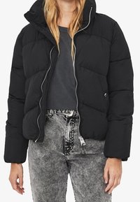 Černá péřová bunda s prošívaným vzorem, kapucí a stříbrným zipem. Nositelná přes šedé tričko a doplněná šedými džínami.