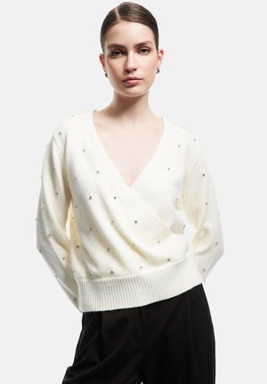 Młoda kobieta z gładkimi włosami, ubrana w biały sweter w serek ozdobiony małymi błyszczącymi koralikami oraz czarne spodnie, na jednolitym tle.