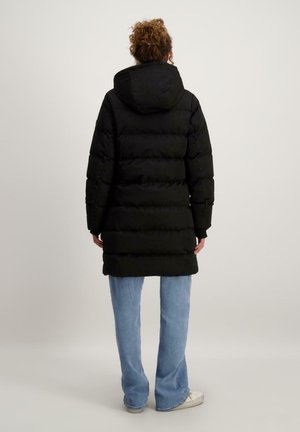 Zwarte pufferjas met een capuchon, voorzien van een doorgestikt ontwerp, lange mouwen en een middellange snit, gedragen over lichtblauwe jeans en witte sneakers.