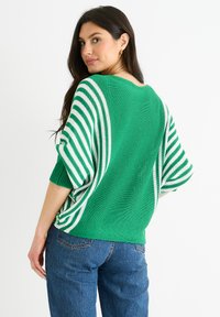 Grüner Strickpullover mit weißen Streifen an den Ärmeln und am Rücken, mit lässigem Schnitt und weitem Ausschnitt. Kombiniert mit blauen Jeans.