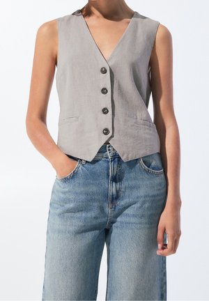 Mouwloos grijs gilet met knopen gecombineerd met lichtblauwe denim jeans met hoge taille, handen in de zakken en neutrale achtergrond.