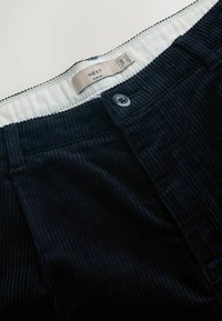 Pantalon en velours côtelé bleu marine au design ajusté, avec une fermeture à bouton et une étiquette indiquant "NEXT". Texture distincte à côtes verticales visible.