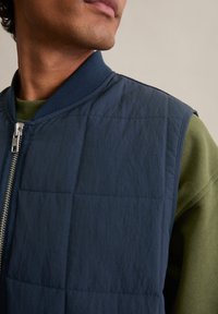 Dunkelblaue gesteppte Weste mit strukturierter Oberfläche, ausgestattet mit einem silbernen Reißverschluss und einem gerippten Kragen. Wird über einem olivgrünen Sweatshirt getragen.