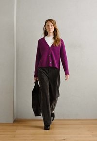 Purple knitted cardigan with a V-neck and buttons, paired with a white turtleneck and gray wide-leg pants. Black shoes and a tote bag.