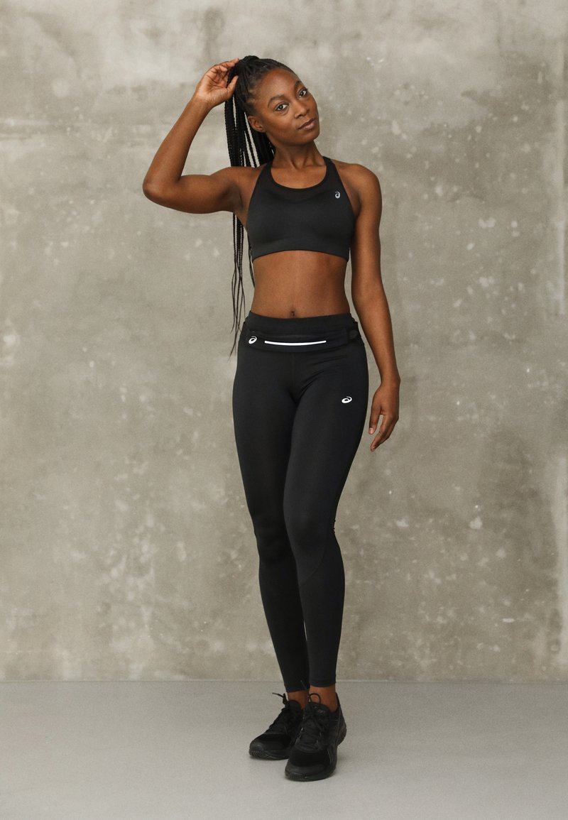 Sutien sportiv negru, cu un design atletico, și colanți cu talie înaltă, ambele realizate dintr-un material neted și elastic cu accente reflectorizante. Încălțămintea este formată din adidași negri.