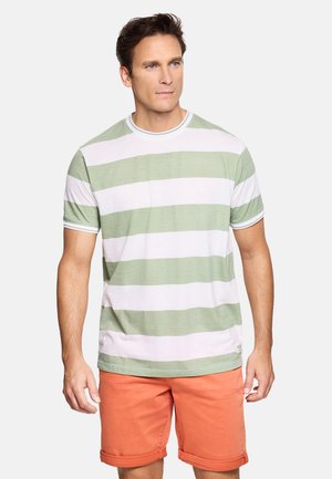 Hombre vistiendo una camisa de manga corta de rayas blancas y verde claro con pantalones cortos naranjas remangados, de pie frente a un fondo liso.