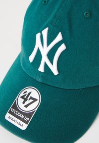 Teal baseball cap with adjustable strap, featuring white embroidered "NY" logo and a black and white tag reading “47 CLEAN UP.” Textured fabric.