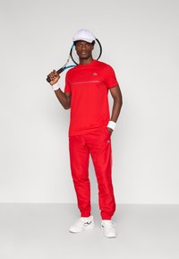 Maglietta sportiva rossa con una striscia orizzontale, pantaloni rossi abbinati, polsini bianchi e un cappellino bianco, mentre tiene una racchetta da tennis.