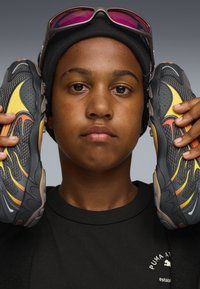 Schwarze und graue Sportschuhe mit gelben und orangefarbenen Akzenten. Das Obermaterial zeigt eine Mesh-Struktur und ein geschwungenes Design, gehalten von einer Person in einem schwarzen Shirt.