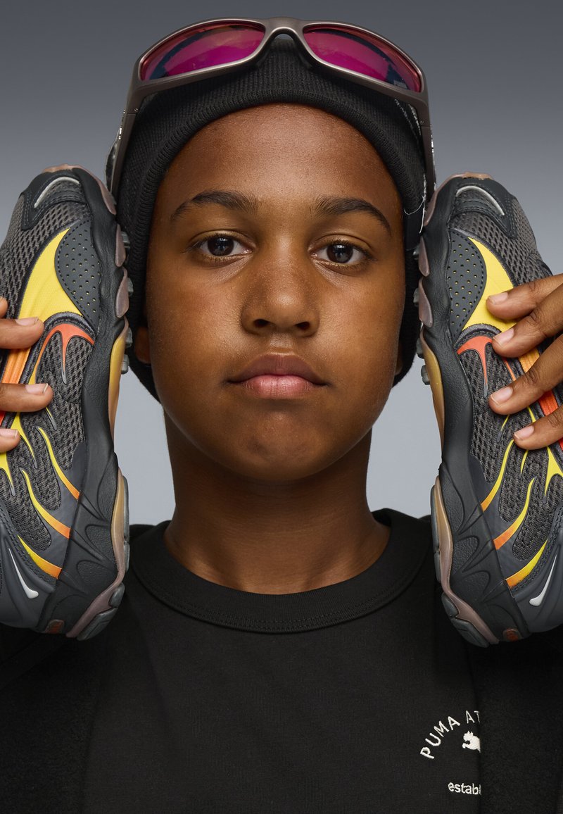 Schwarze und graue Sportschuhe mit gelben und orangefarbenen Akzenten. Das Obermaterial zeigt eine Mesh-Struktur und ein geschwungenes Design, gehalten von einer Person in einem schwarzen Shirt.