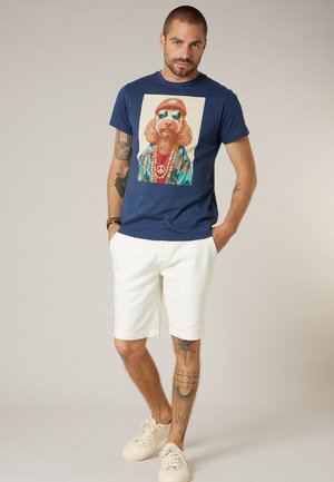 Homme portant un t-shirt bleu marine avec un motif coloré de chien, un short blanc, des baskets blanches, et plusieurs tatouages sur les bras et les jambes, debout de manière décontractée.