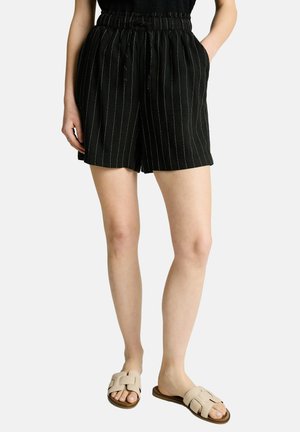 Vrouw die zwarte shorts met fijne strepen en elastische taille draagt, handen in de zakken, en beige slip-on sandalen, afgebeeld van taille tot voeten.