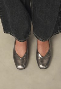 Bailarinas plateadas metálicas con puntera afilada y textura suave, colocadas sobre unos vaqueros oscuros rematados en los tobillos.