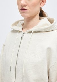 Γυναίκα που φοράει ζακέτα με φερμουάρ σε ανοιχτό μπεζ χρώμα, με κορδόνια και διακριτικό λογότυπο Calvin Klein στο στήθος, σε απλό λευκό φόντο.