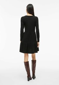 Femme aux longs cheveux bruns portant une robe en maille noire longueur genoux et des bottes brunes à talons hautes jusqu'aux genoux, se tenant contre un fond blanc.