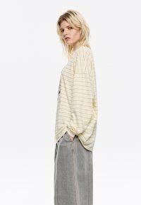 Donna bionda in un top cremoso a maniche lunghe e larghe con strisce nere e pantaloni grigi a gamba larga, in piedi di lato con le mani in tasca.